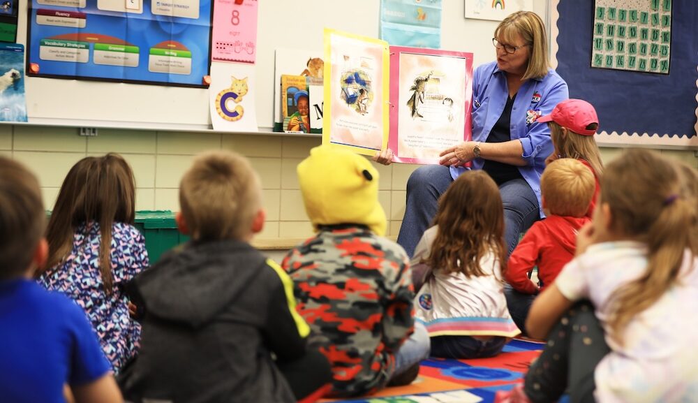 A woman sitting on a small chair reading to children in a classroom.