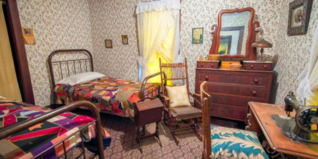 A small bedroom with two antique beds, dresser, and nightstands, featuring a cozy, multicolored knit bedspread and patterned curtains.