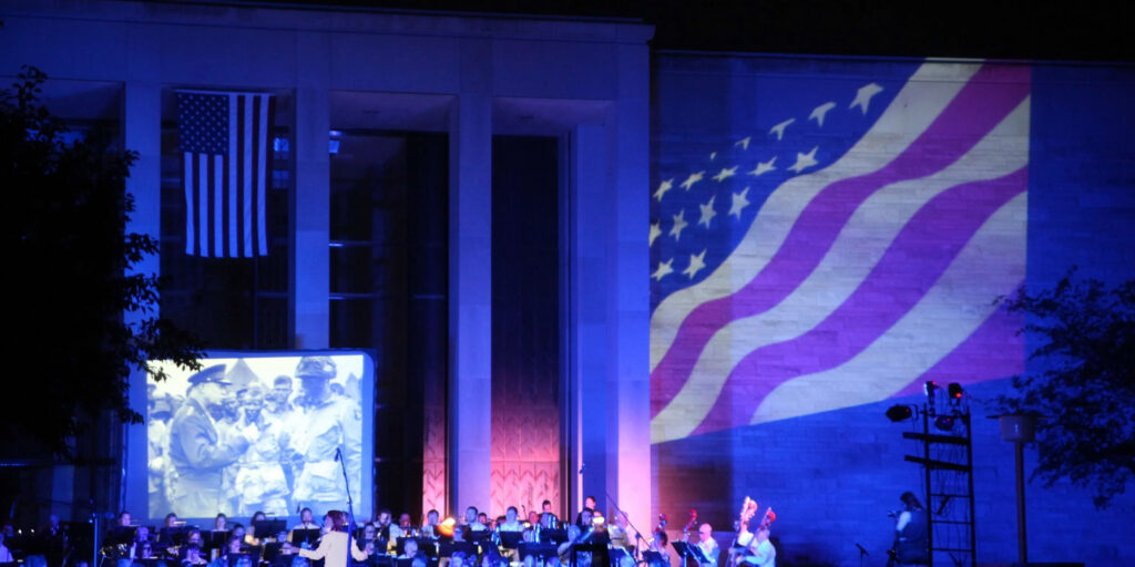 A flag display with a building. There is a group of people in the background, playing instruments.