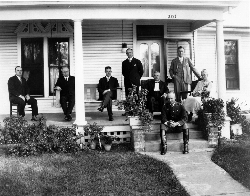 A photograph captures the Dwight D. Eisenhower family gathered for a reunion at his boyhood home in Abilene. They are posing on the front porch of the house, all dressed in semi-formal attire, smiling and facing the camera.