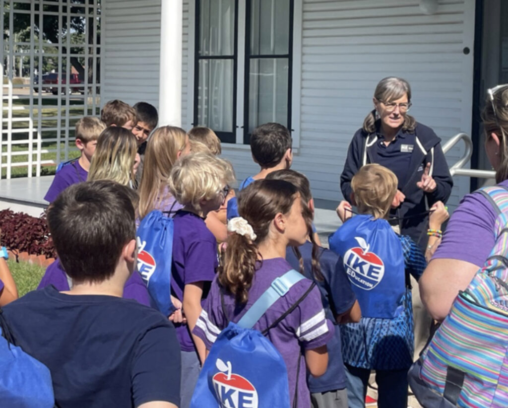 A woman in purple shirt stands infront of a group of children, who are gathered around her. They are outside infront of a building with columns and a white fence. The woman is gesturing with her hands.