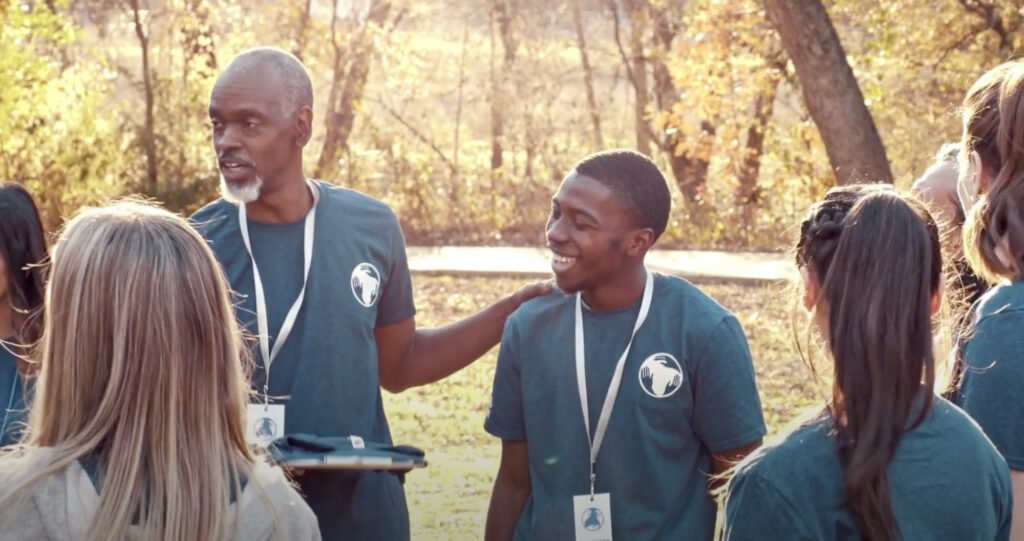 The image shows a group of people in an outdoor setting. The central figure seems to talk to the group, while the group seems to listen. They wear matching shirts and lanyards.