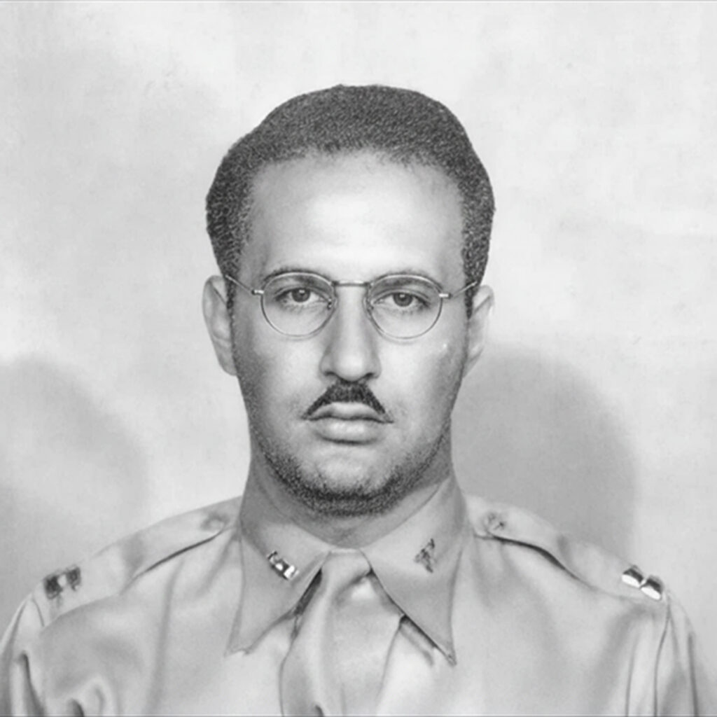 A sepia photograph of a man in military uniform looking serious. He has mustache and wears glasses.