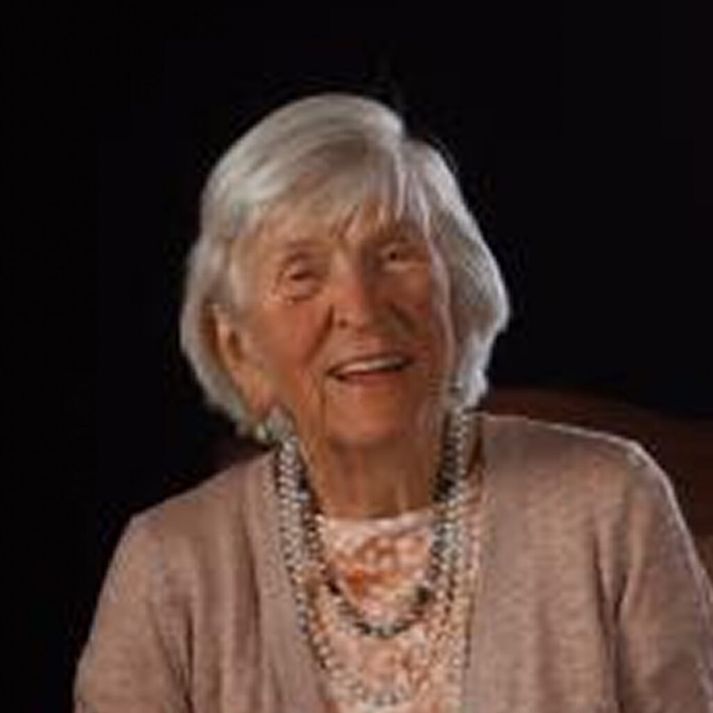 The image is of an elderly woman smiling at the camera. She has short gray hair, and her skin appears to be fair with a few wrinkles. She's wearing a necklace around her neck. There are no visible texts. The background is dark, which helps to focus on the woman.