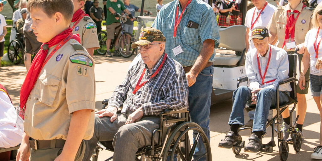 A group of veterans on a city street, some standing, others in wheelchairs. There are also some scouts in uniform, wheeling the veterans.