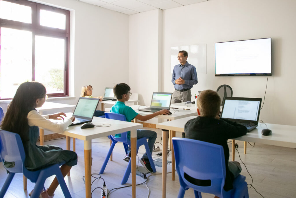 A teacher stands in front of a group of children seated at desks with laptops, engaging in an interactive lesson.