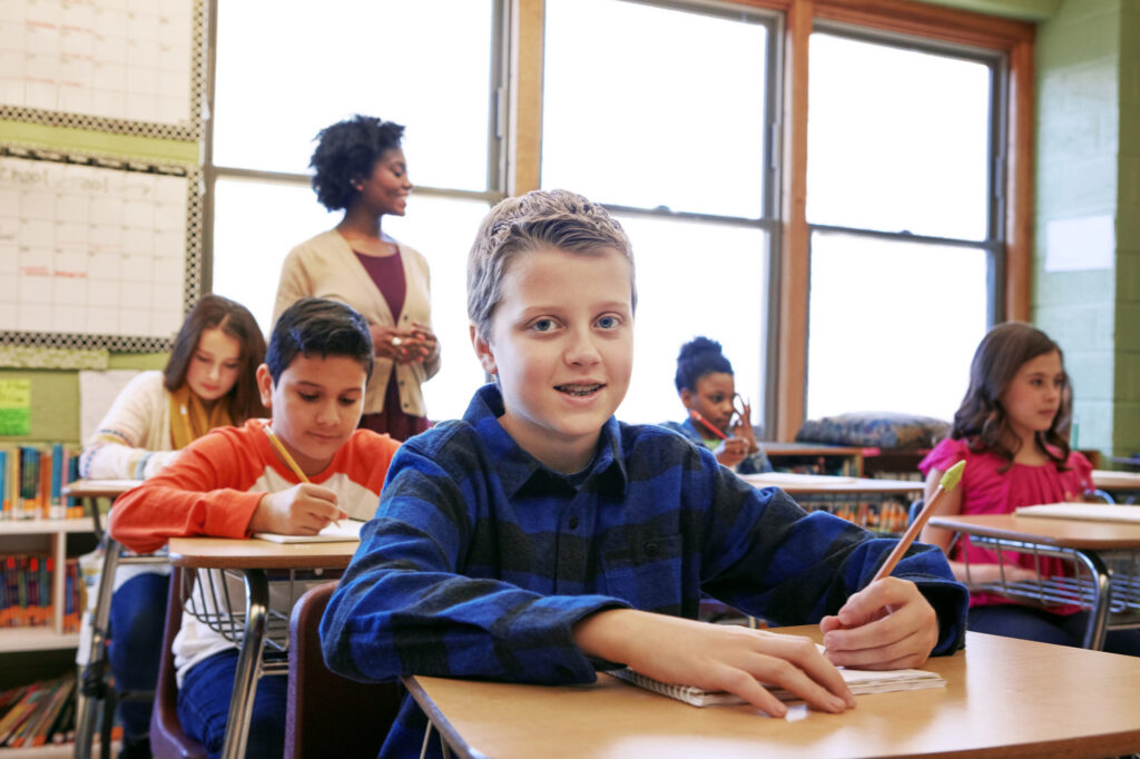 A student sitting at a desk in a classroom, holding a pencil and paper, with other students at desks behind him. A teacher is standing by the window. The room has natural light from the windows on the left side of the image.