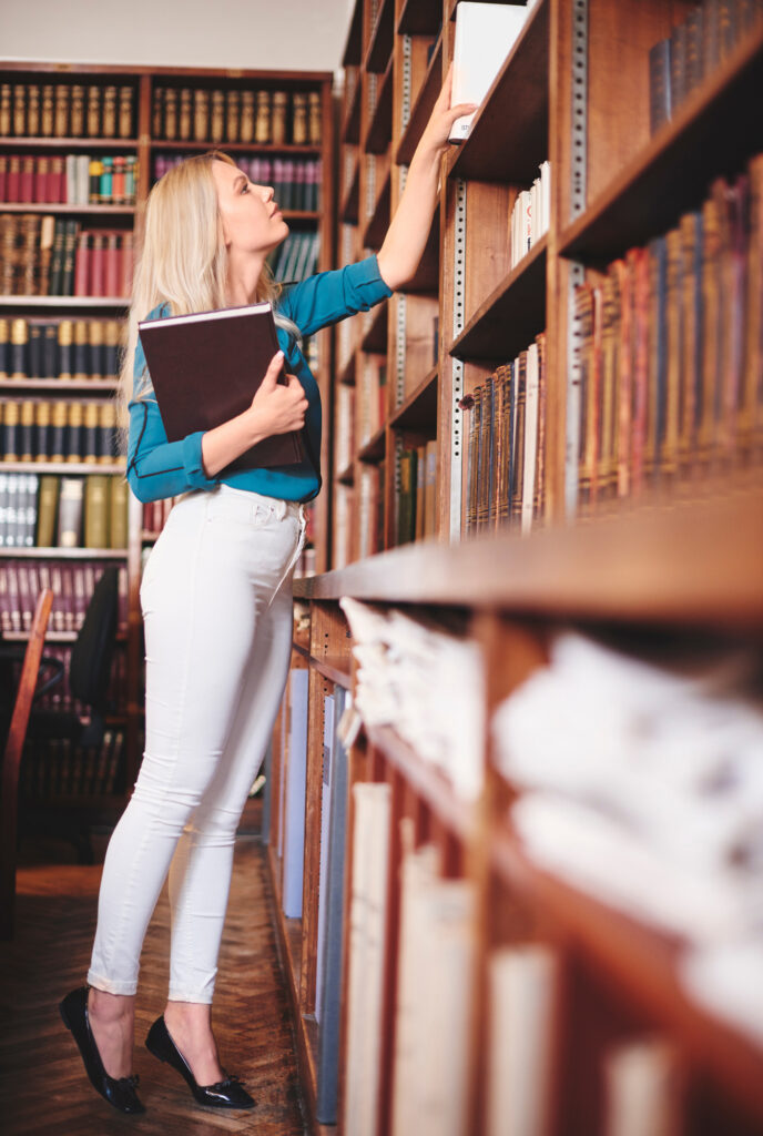 Woman in blue shirt reaching for book on shelf.