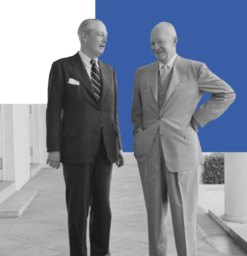 Two men in suits standing together. One man is smiling and leaning forward slightly. The man to his right stands with arms crossed. They appear to be in an outdoor setting, possibly on a balcony or patio.