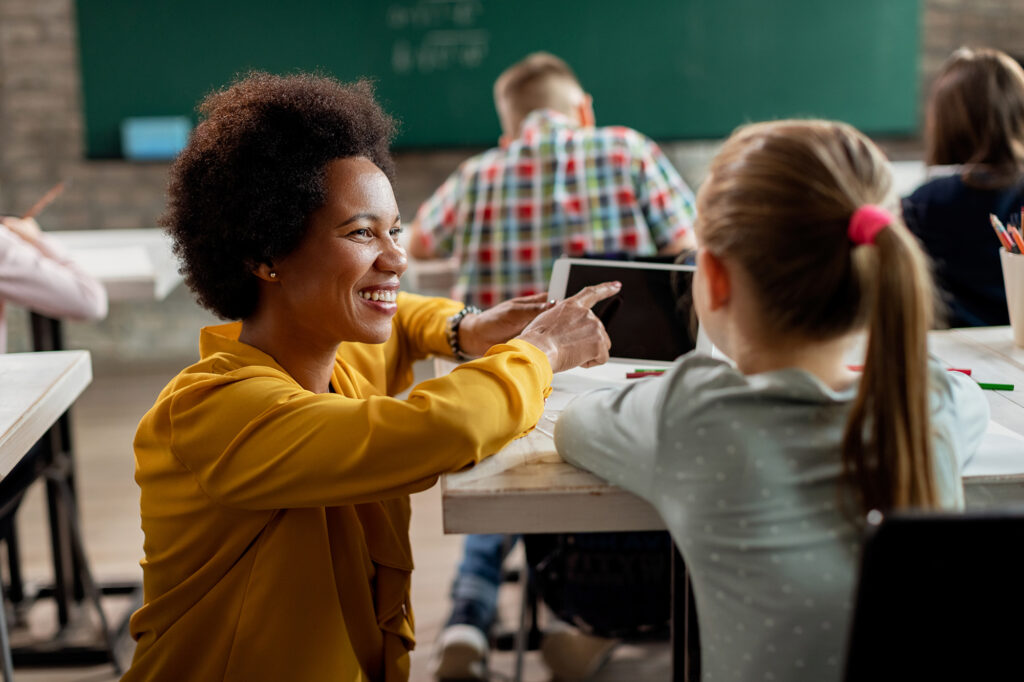 This is an image of a woman teaching a class of children. The teacher is crouched beside a student and is pointing at a tablet in her hand. She has a broad smile on her face. There are several students seated at desks, engaged in what seems like a learning activity.