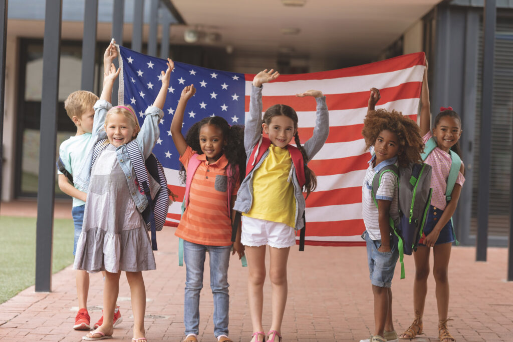 Young students, some wearing backpacks, proudly hold up an American flag as they pose for a photo in front of a school building.