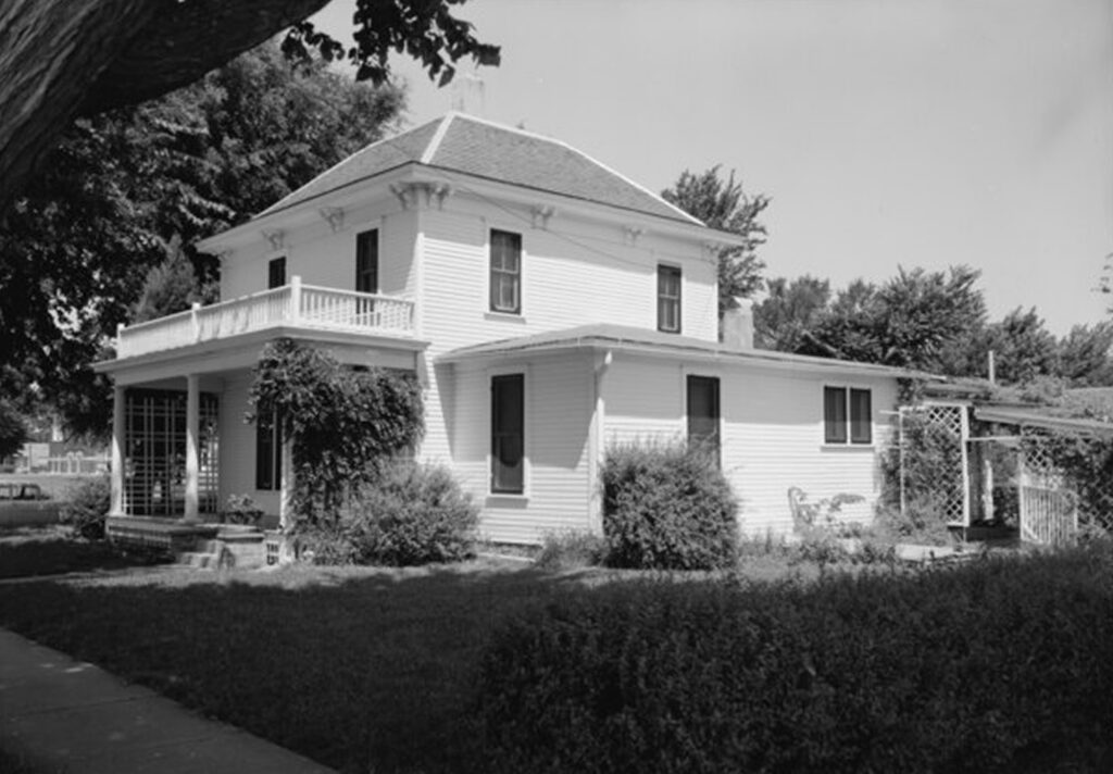 A black and white photograph of an old two-story house with a pitched roof, multiple gables, and a porch with columns at the front. The house has a large window on the second floor and a chimney. It is situated in a landscaped yard with trees and bushes surrounding it.