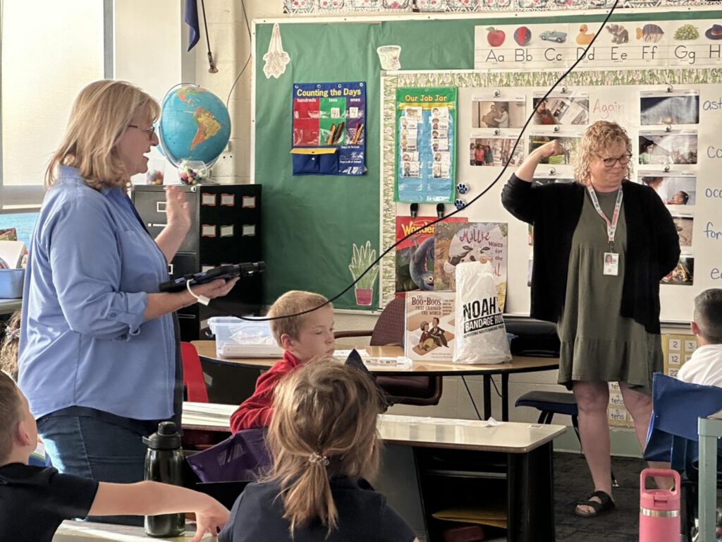 Two women and a groupd of children are in a classroom. The children are at desks.