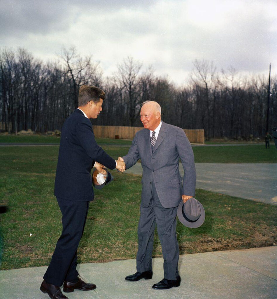 This photograph depicts a meeting between President John F. Kennedy and former President Dwight D. Eisenhower. They are shaking hands in an outdoor setting.