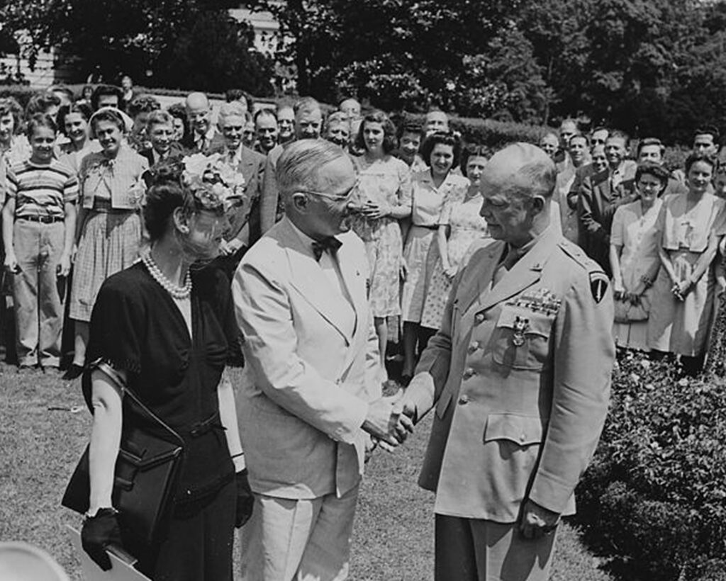 The image shows an outdoor gathering where two men are shaking hands. In the background, there is a group of people looking towards the three central figures. The individuals appear to be engaged in a formal interaction or conversation. One of the men is Dwight D. Eisenhower.