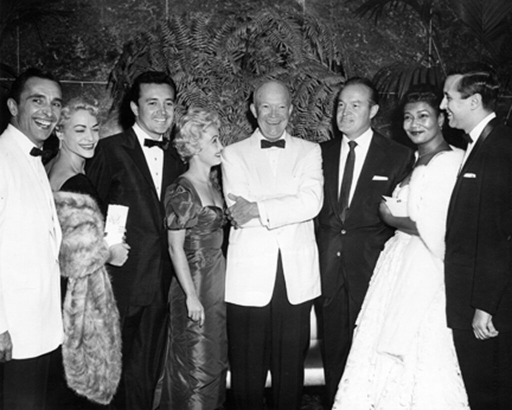 The image is a black and white photograph featuring a group of seven individuals in formal attire standing together. They are posing for a picture with smiles on their faces. The setting suggests a formal event or gathering.