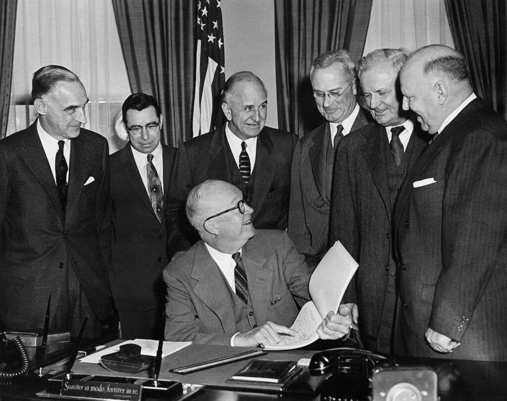 A black-and-white photograph of a group of men in suits gathered at a desk with documents; one man signing a paper while others look on.