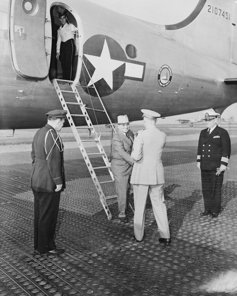A military airplane on tarmac, opening the side door to reveal the interior; men in uniforms stand and greet each other.