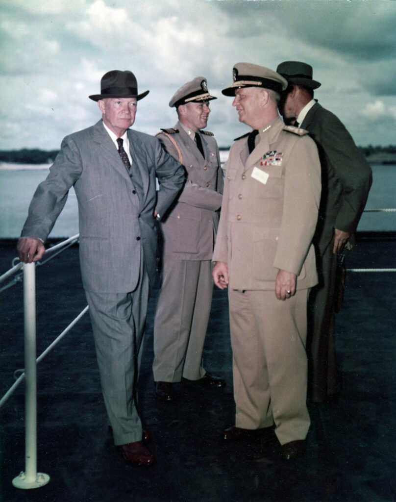 Four men on a boat deck; two are wearing suits and ties, and the others are wearing military uniforms; they seem to be engaged in conversation.