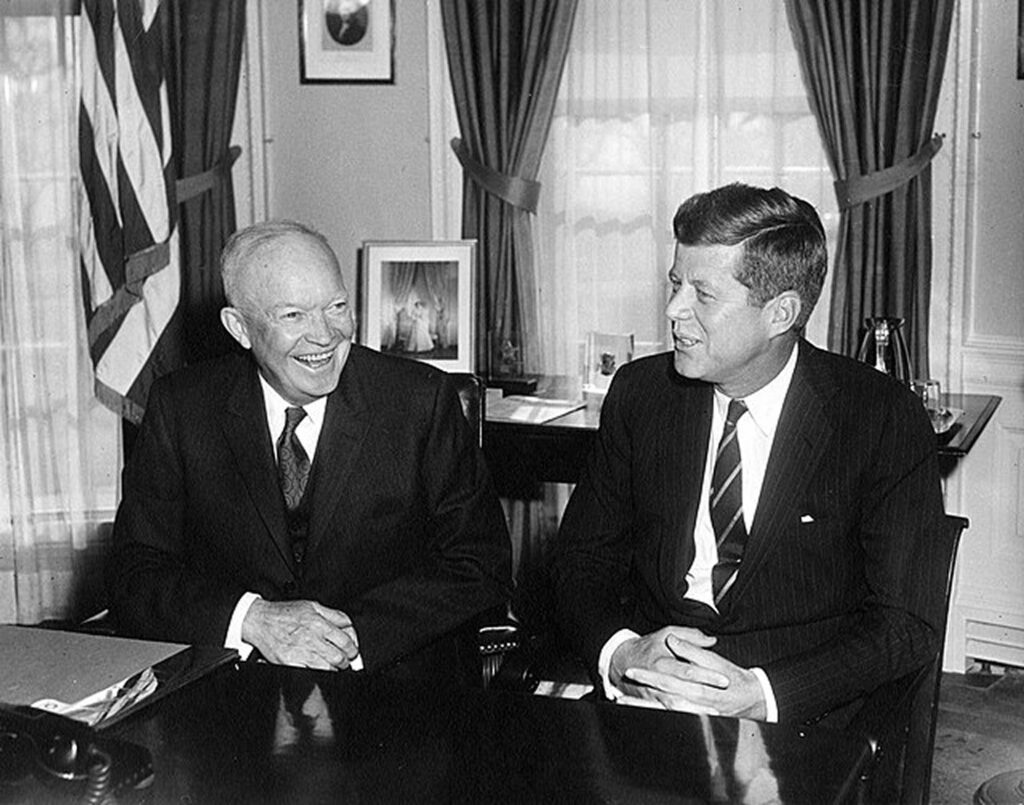 A black-and-white photograph of a meeting between outgoing President Dwight D. Eisenhower and President-elect John F. Kennedy. They are seated at a desk, smiling and engaged in conversation.