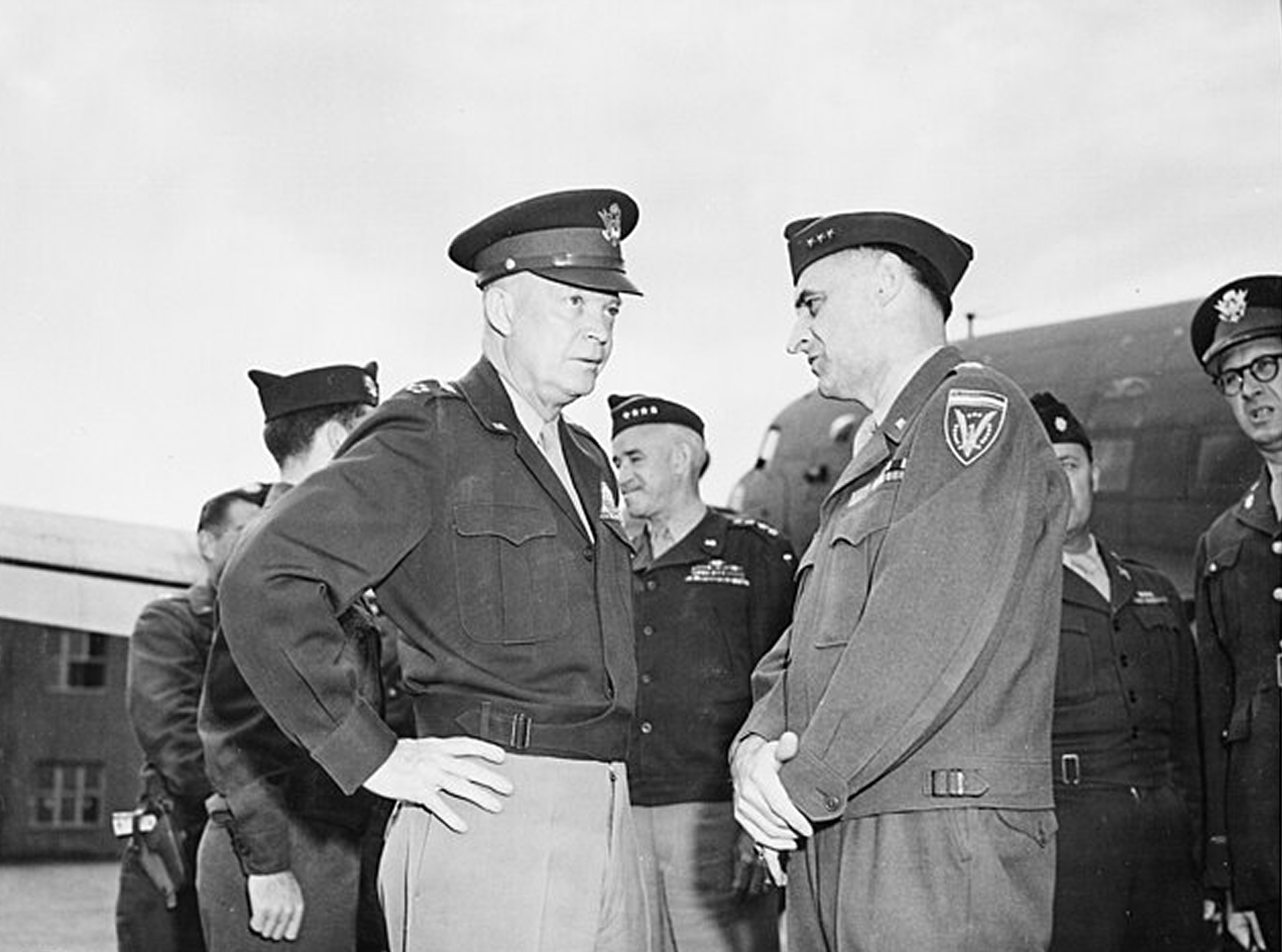 Dwight D. Eisenhower in conversation with another man on an airfield. There are men in uniform behind them.
