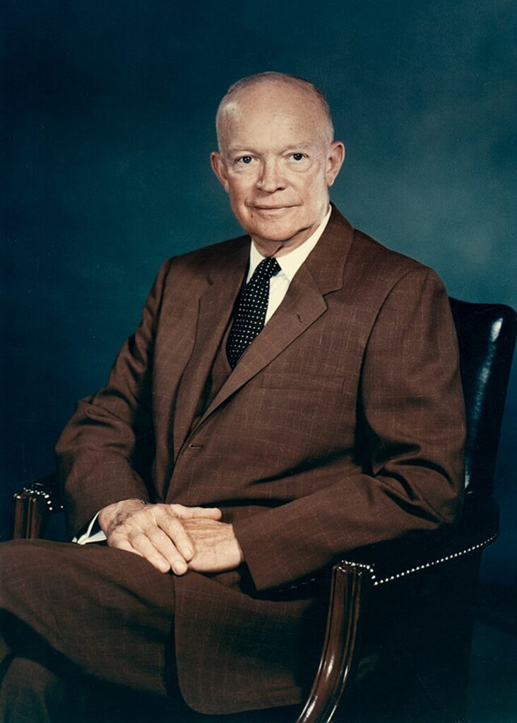 The image shows Dwight D. Eisenhower seated in a chair. He is wearing a dark suit and tie, suggesting a formal attire, possibly for an official portrait. His posture is relaxed yet dignified, with his arms resting on his lap.