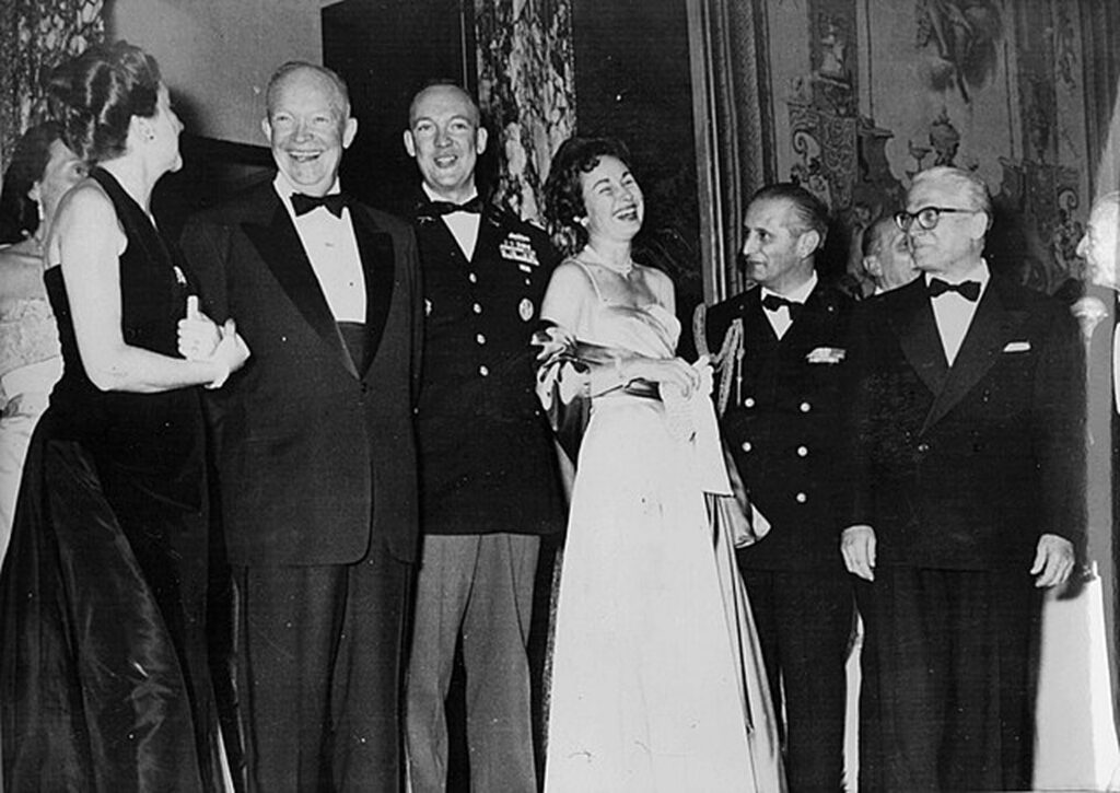 A vintage black and white photograph captures a formal event, featuring several men and women in evening attire. The group is posing for the camera with smiles. Some individuals are wearing ties, and the women are in dresses. They stand in front of a wall adorned with decorative elements.