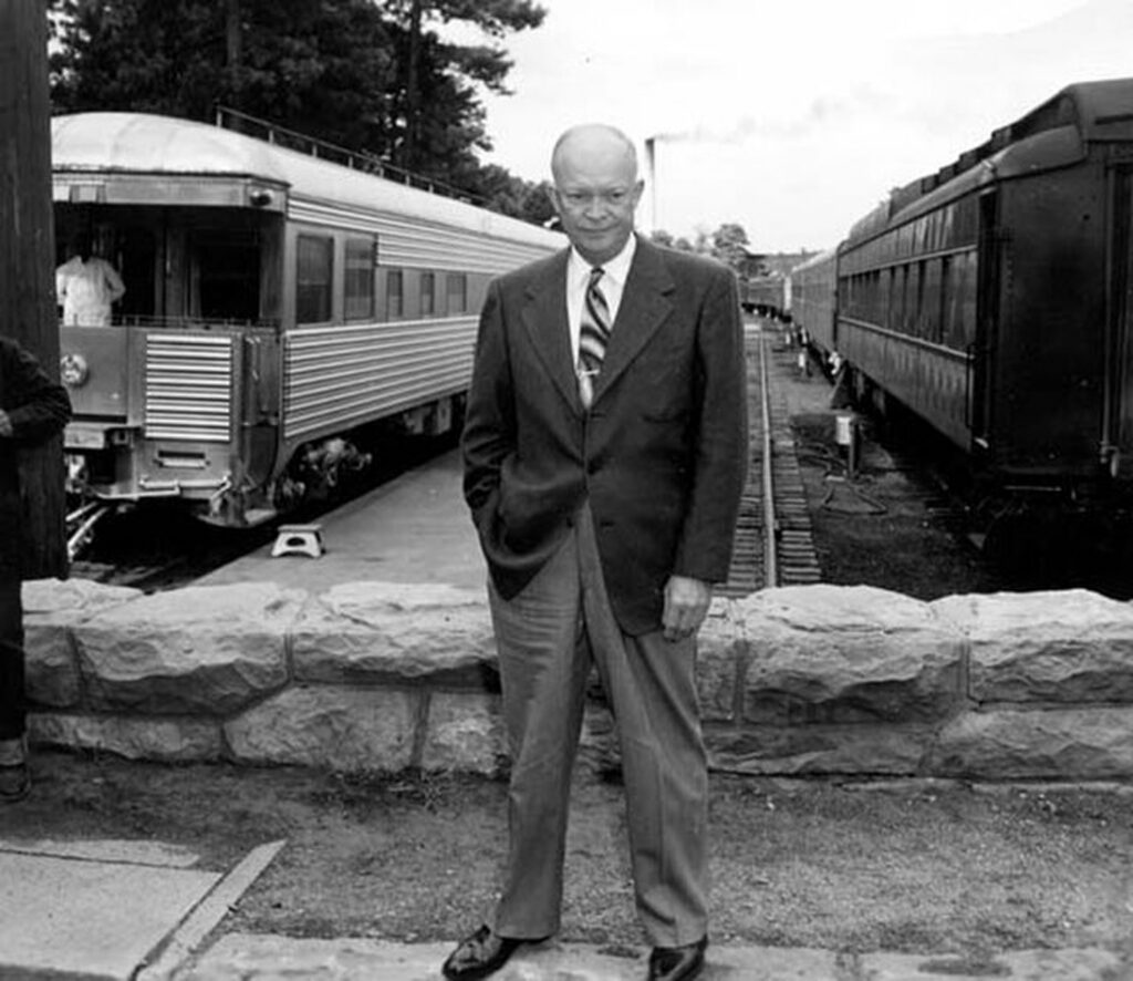 This is an old photograph featuring Eisenhower. He's wearing a dark suit and tie. Behind him is a vintage silver passenger train with several cars visible, parked near what appears to be a platform. The setting suggests an old-fashioned train station