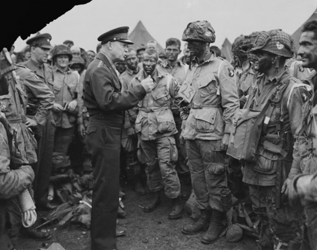 Military men in field uniforms gathered around a man standing and addressing them. He is distinguished by his formal attire with insignia and appears to be giving orders or instructions. Their attention is focused on him. They are outdoors, possibly in an army encampment or training area.