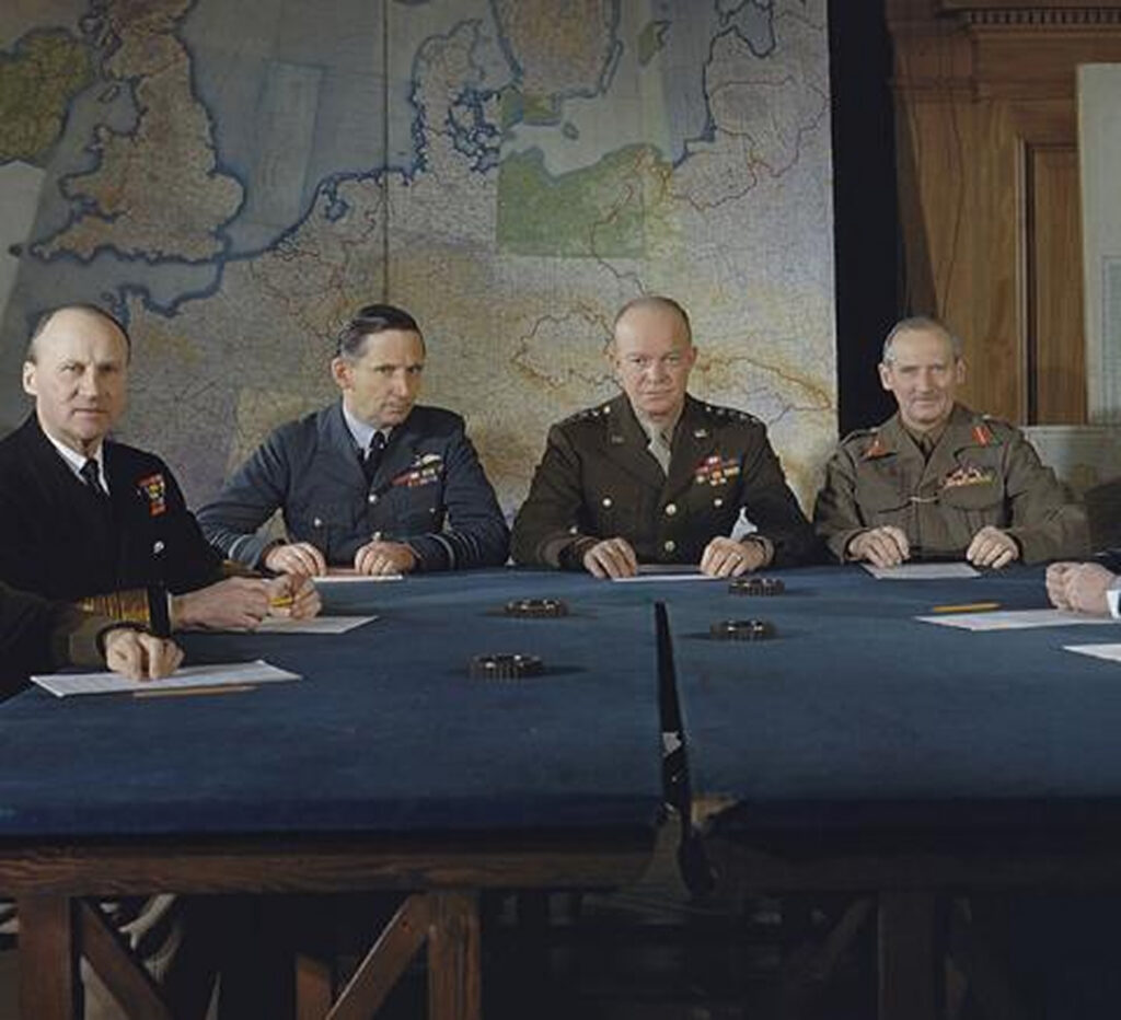 Five men in military uniforms are seated around a conference table. They have papers and pens in front of them, and are looking at the camera.