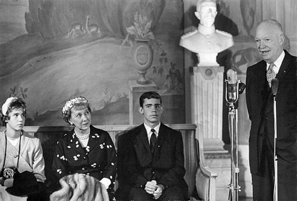 A group of people on stage at what appears to be a formal event. A central figure is speaking into a microphone and wearing a suit with a tie. To the left, there are three individuals sitting, attentively listening to the speaker.
