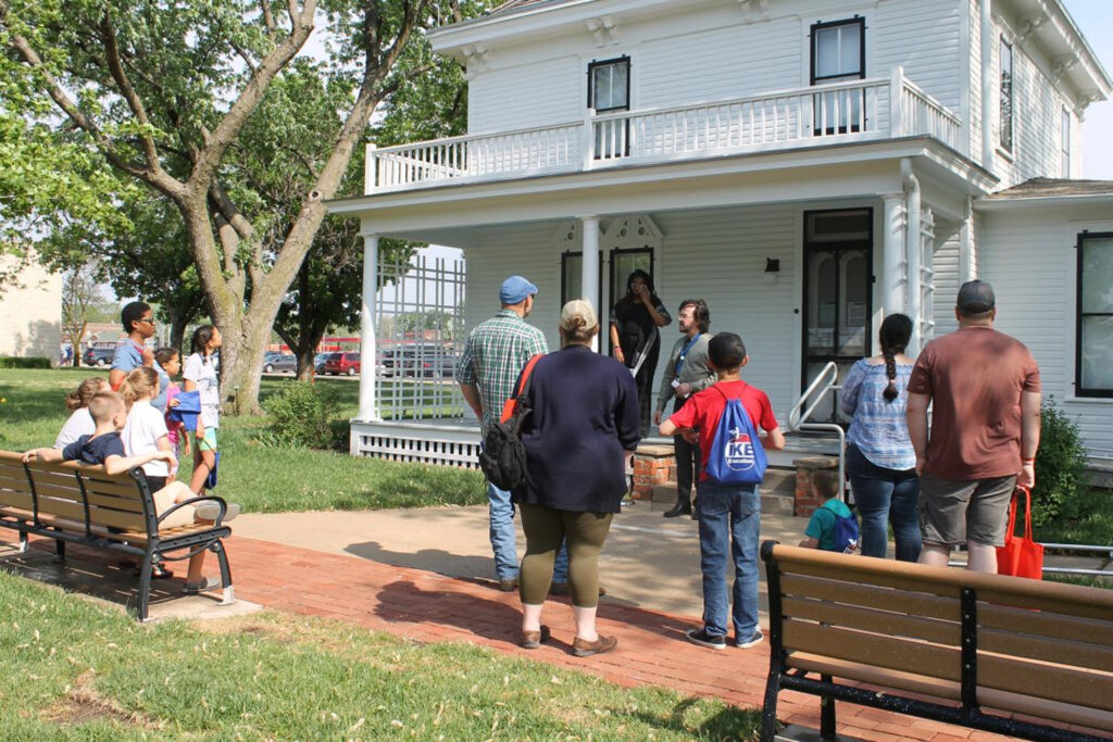 A group of people standing in front of a white two-story house with a wraparound porch. They seem to be gathered for an event or meeting. One person is standing in front of the porch seems to be addressing the people. The setting suggests a residential neighborhood, with trees lining the street.