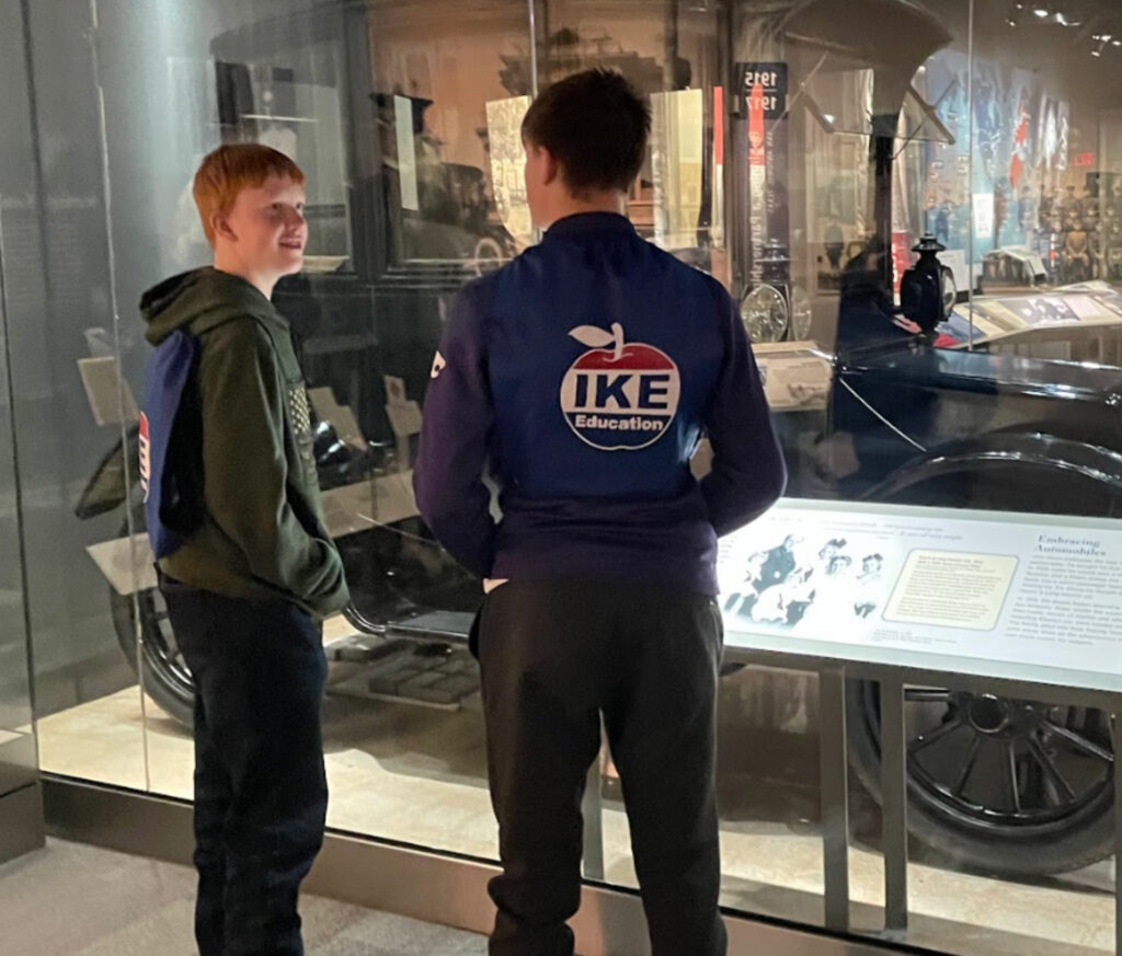 Two boys in front of a vintage car exhibit. Both boys have their hands in their pockets, reading a placard. Both are wearing jackets.