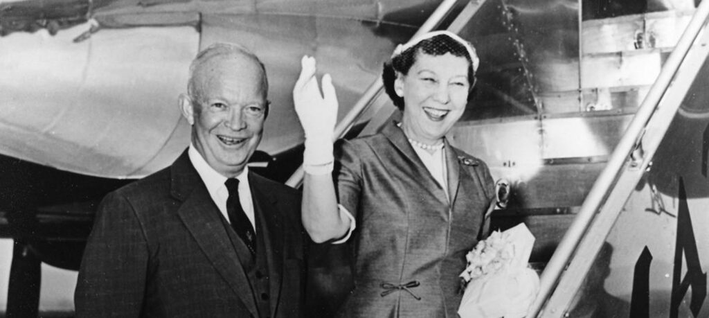 A black-and-white photograph capturing two people in formal attire waving from an airplane.
