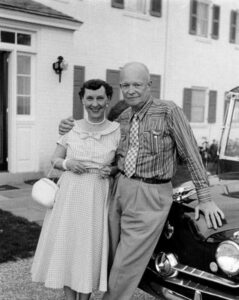 Mamie and Dwight D. Eisenhower posing together in front of a classic car, with an affectionate side hug. The woman wears a dress with a full skirt, and they both appear happy and content.