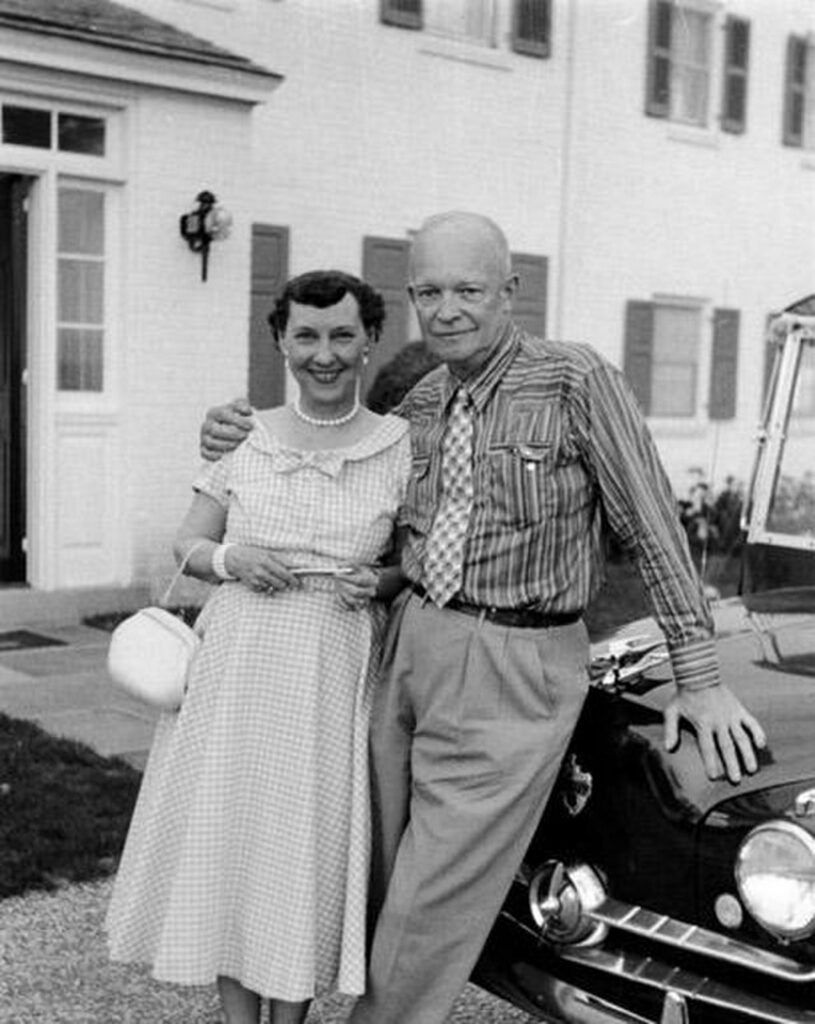 Mamie and Dwight D. Eisenhower posing together in front of a classic car, with an affectionate side hug. The woman wears a dress with a full skirt, and they both appear happy and content.