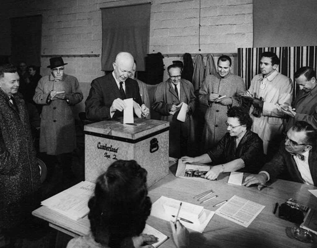 In black and white, a group of people gather around Dwight D. Eisenhower who appears to be casting a vote; he is placing it into a box.
