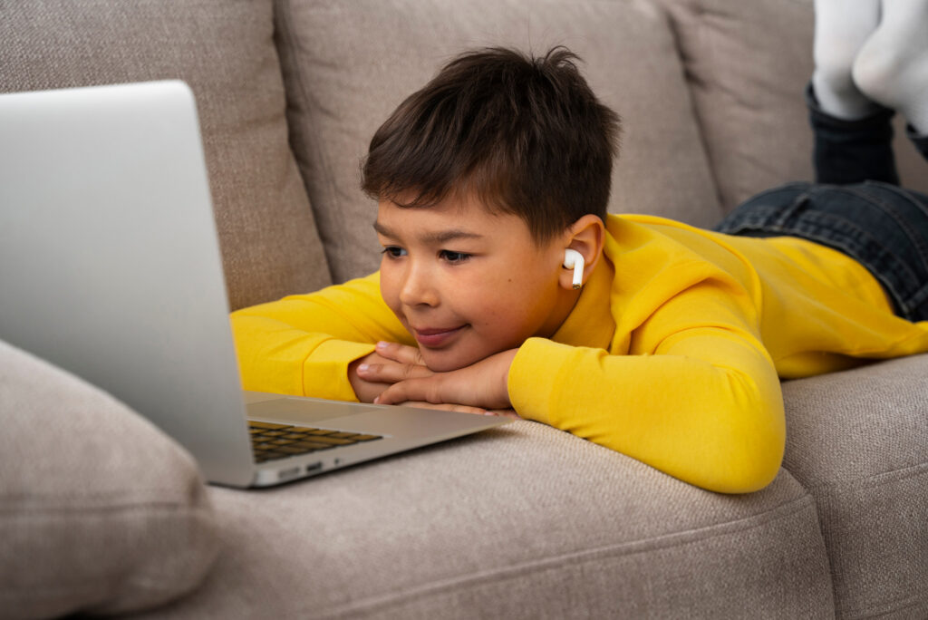 A young boy lying on a couch with his feet up, engrossed in using a laptop computer. He has short dark hair and is wearing a yellow shirt.
