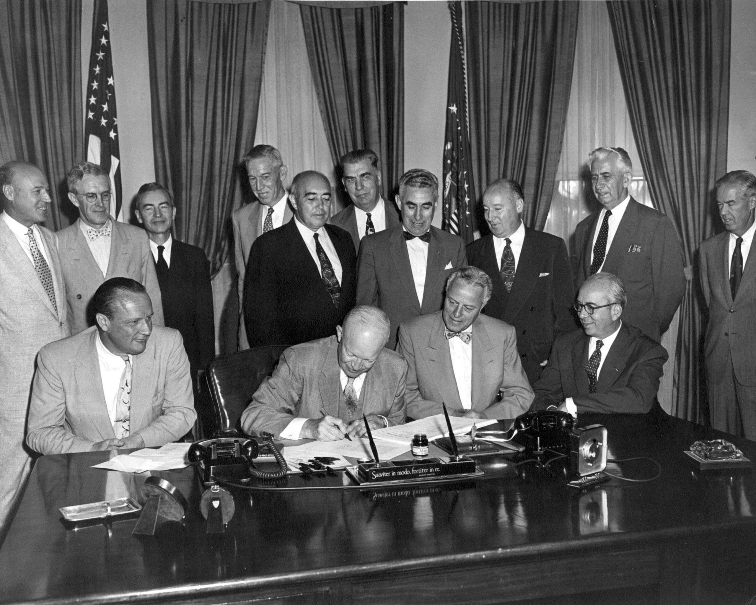 Politicians gather in formal attire around Dwight D. Eisenhower, who is signing a document at a conference table.