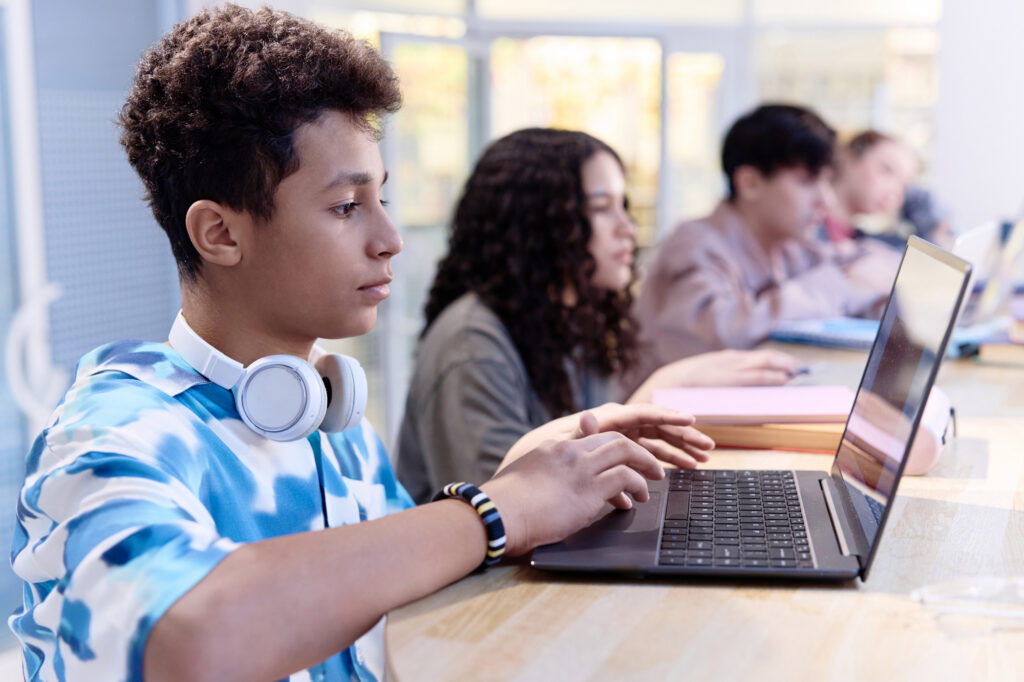 A young boy using a laptop while sitting at a table in an indoor setting with other students around him.