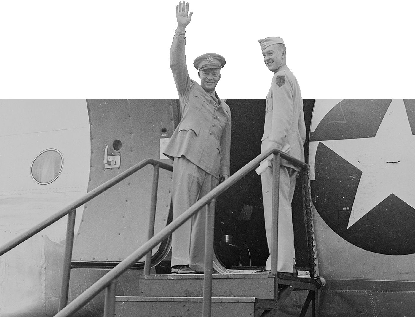Two individuals on steps in front of an airplane. One person is waving, and both are wearing military uniforms with medals.