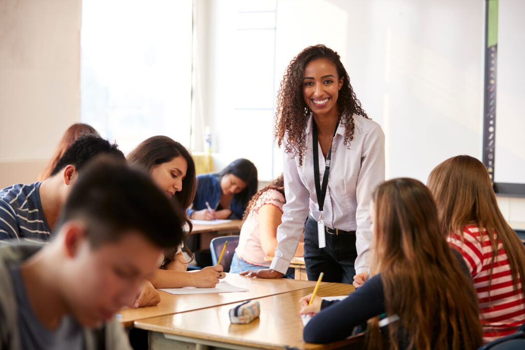 A teacher stands in front of students seated at desks in a classroom. She appears to be leading or instructing the class and is facing forward while smiling at the camera.