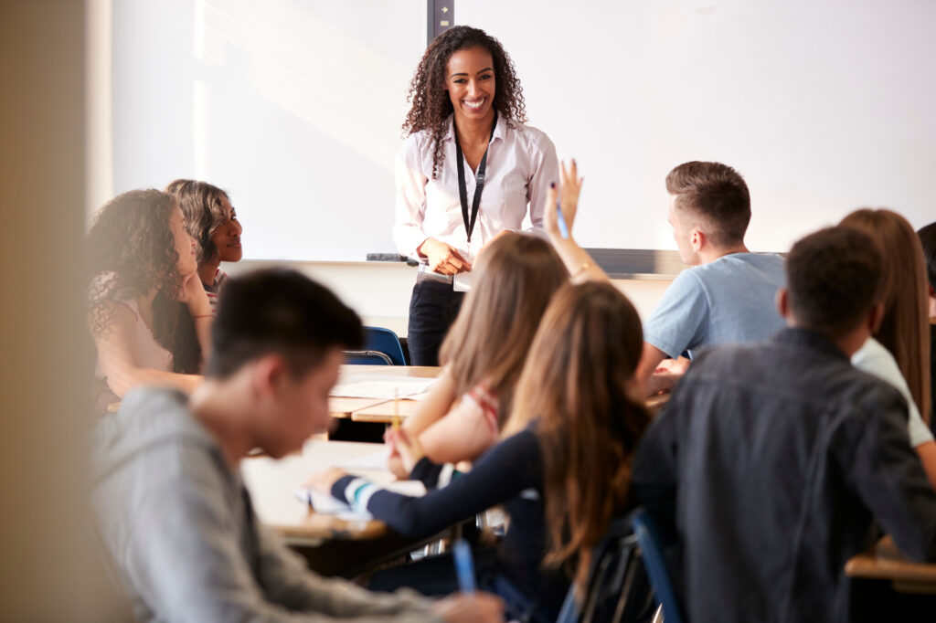A classroom scene with students seated at desks; a teacher in front stands, addressing the class. She wears a white shirt and smiles broadly.