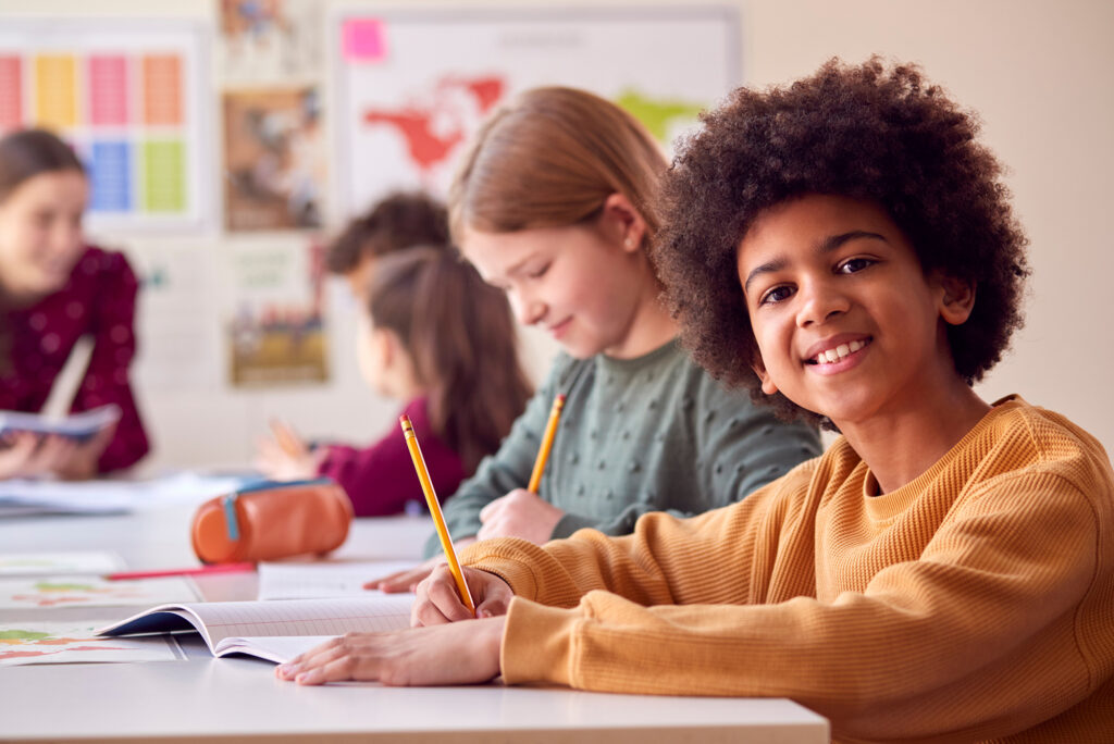 A child sitting at desk in classroom with pencil and book, smiling at camera. Fellow students are in the background, also studying.
