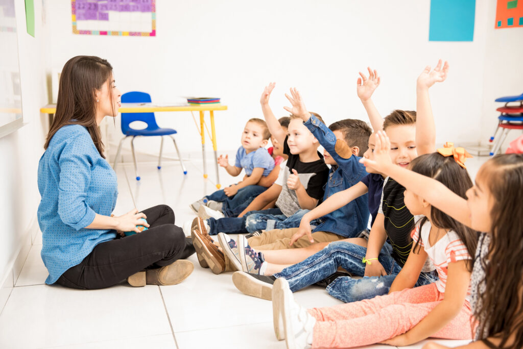 Teacher instructing students in a classroom. Many of the young students are raising their hands while smiling.