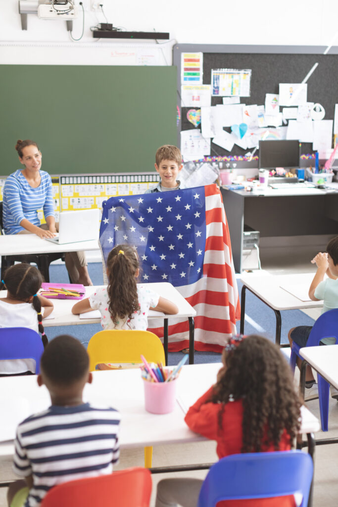 In classroom setting, teachers present, a child holds an American flag in front of the rest of his class.