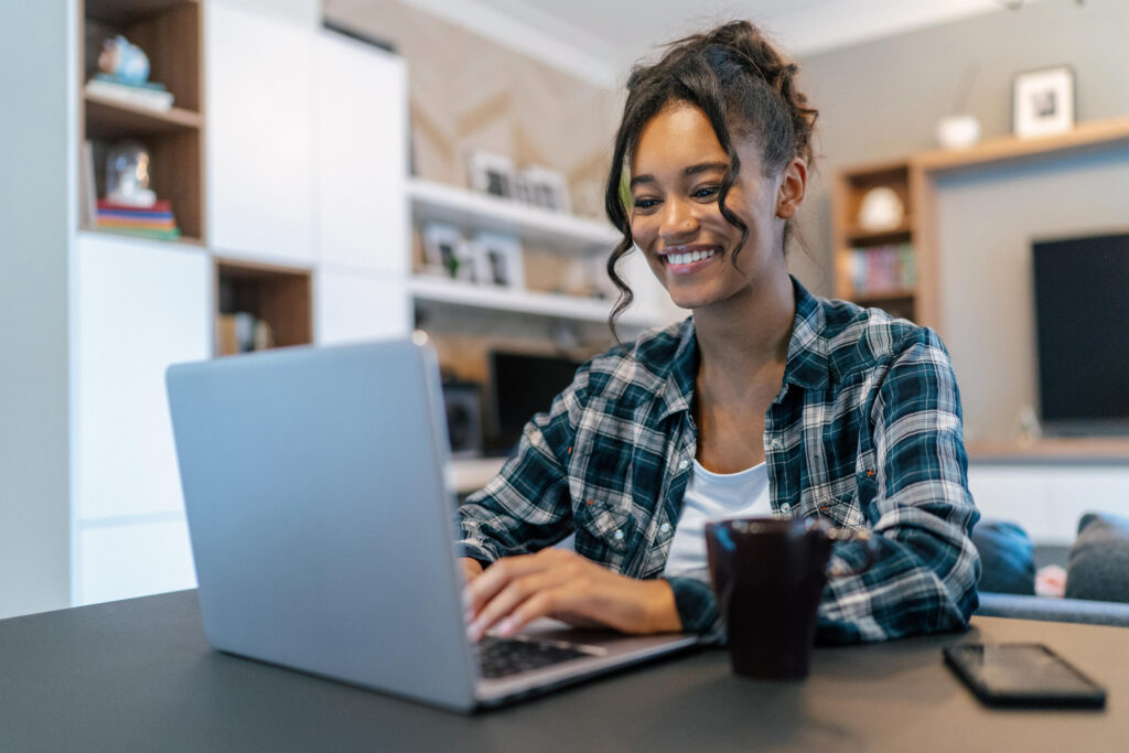 A young woman sitting at a table, using a laptop, smiling.