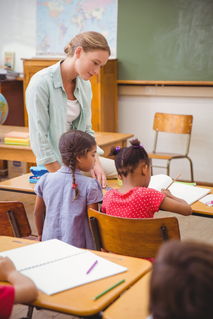 A teacher helps two students at school desks.