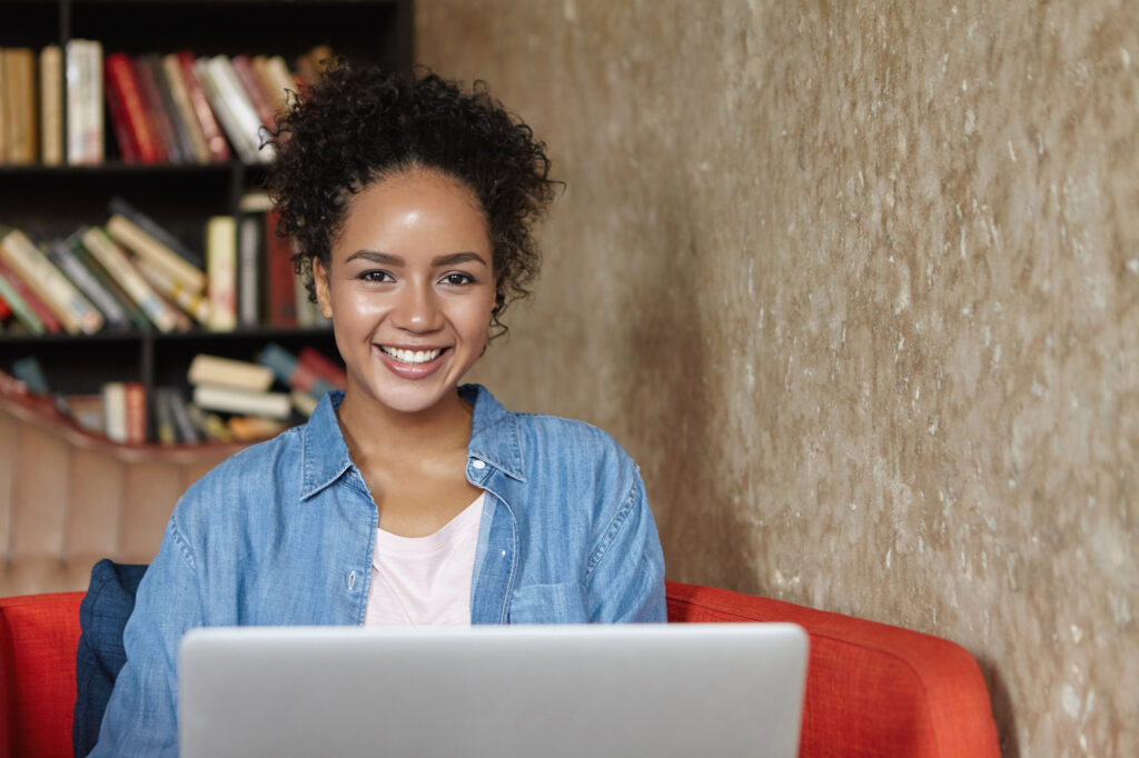 A woman sitting on a red couch, smiling and looking at her laptop computer, which is open and placed on her lap. She has dark hair and is wearing a blue top.
