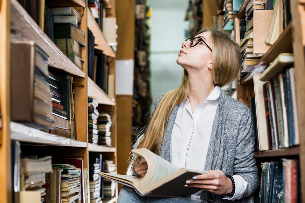 A woman in library, reading book, smiling, surrounded by bookshelves.